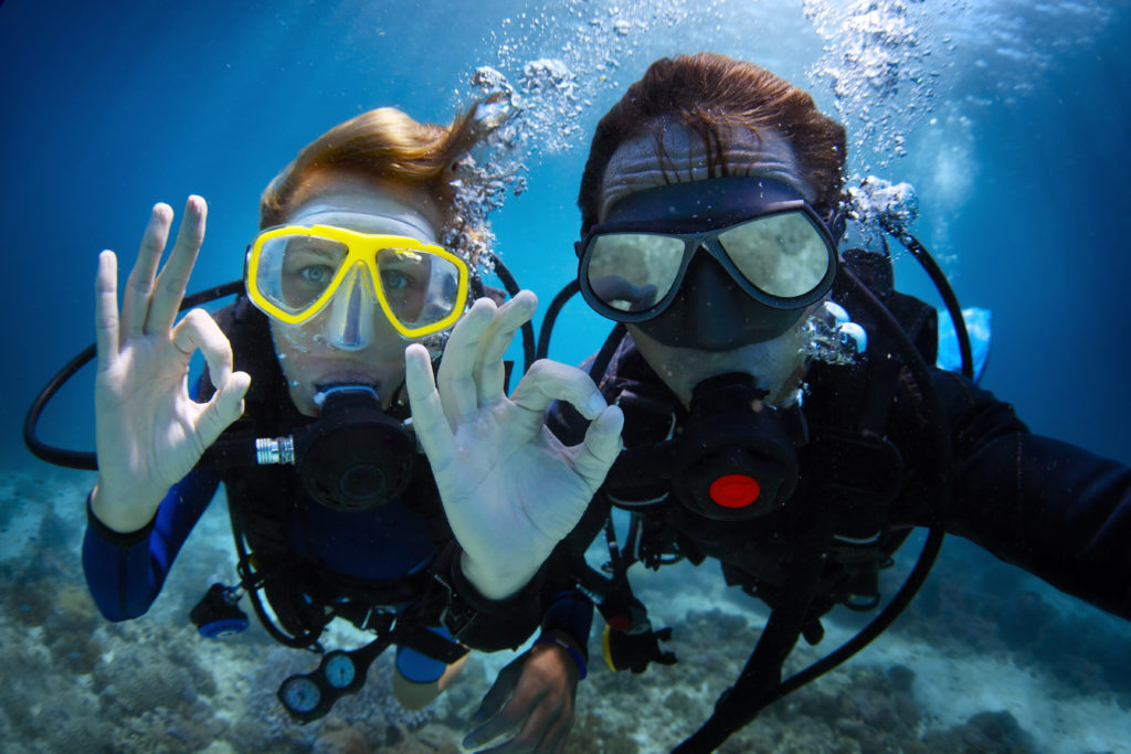 Two men posing underwater with a 'super' hand gesture while scuba diving. The image captures a playful moment during the dive
